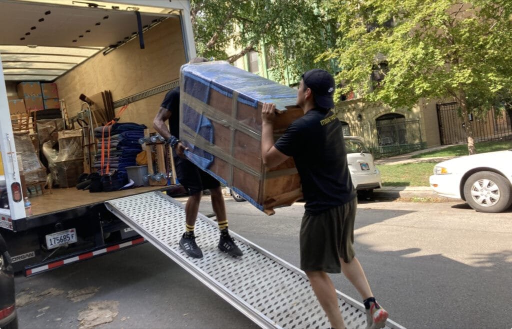 Two men wearing work gloves and safety vests are loading large cardboard boxes into a white delivery truck. One man stands on the ground lifting a box while the other, positioned inside the truck, reaches out to receive it. They appear to be coordinating their efforts in a sunny, outdoor setting with a few trees visible in the background.