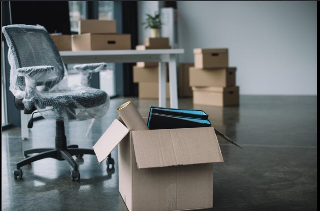 I“A team of movers carefully handling packed boxes and wrapped office furniture in a modern office space, preparing for an organized office move. The office is partially empty with desks, chairs, and computers wrapped in protective materials.”